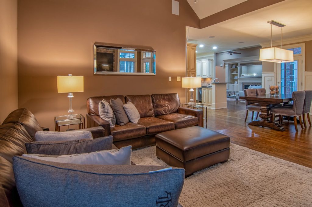 Modern open-plan living room with a large neutral area rug, leather sofa, ottoman, and hardwood floors connecting to dining and kitchen areas