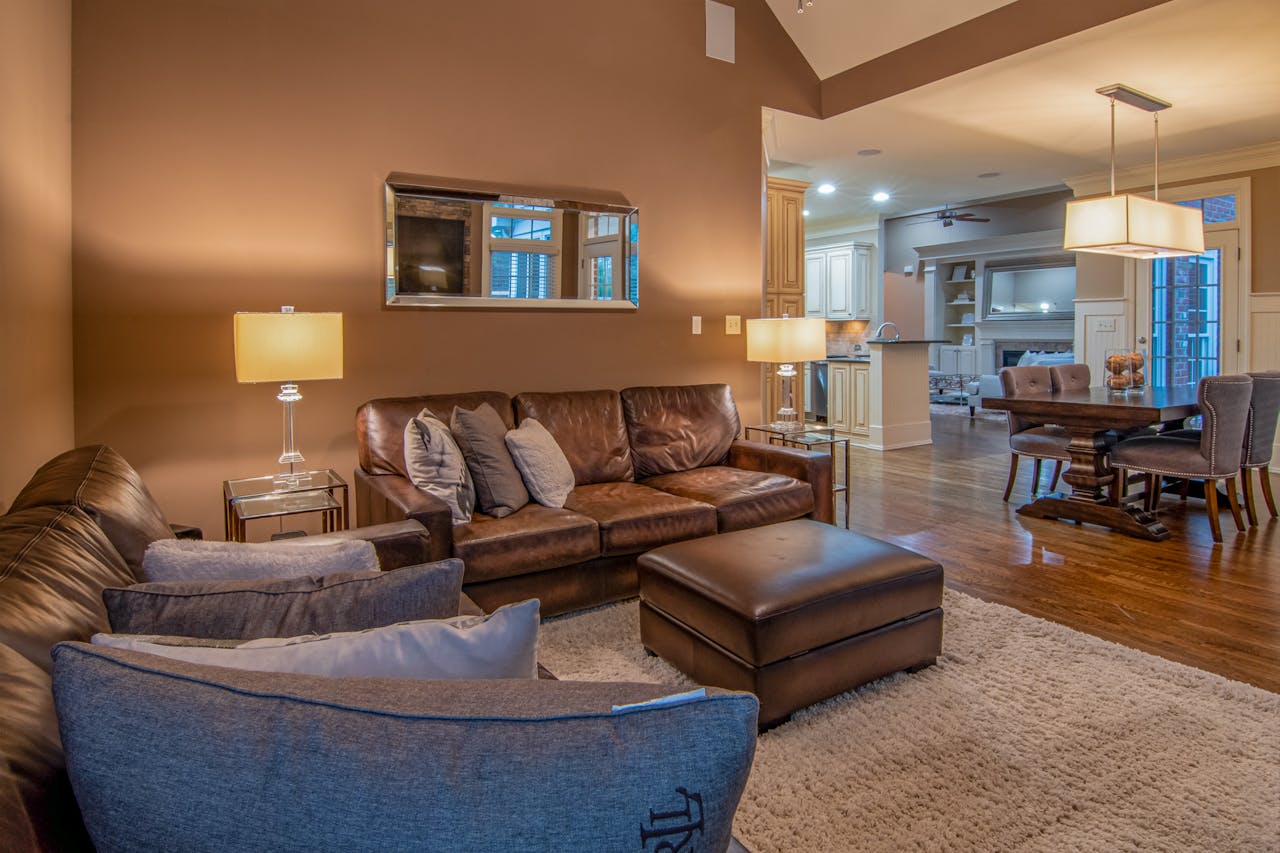 Modern open-plan living room with a large neutral area rug, leather sofa, ottoman, and hardwood floors connecting to dining and kitchen areas